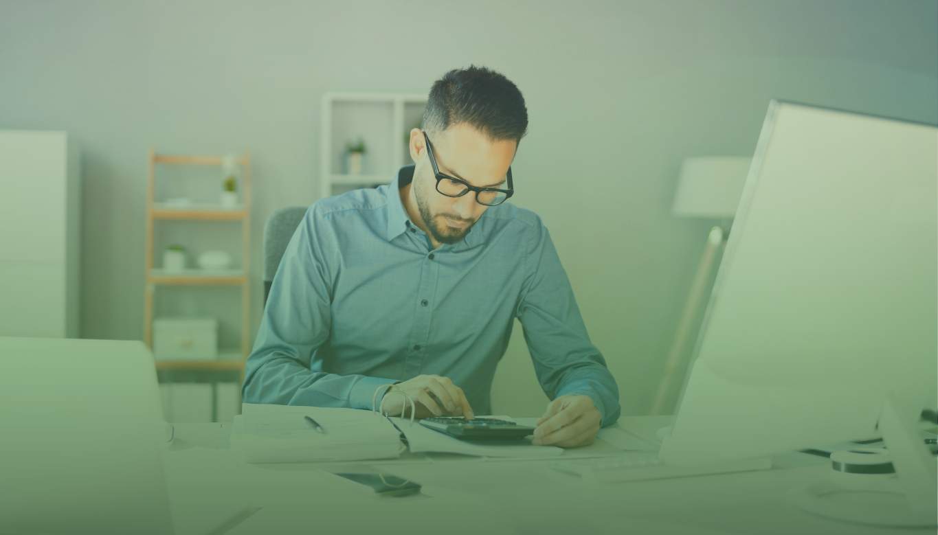 Business owner doing bookkeeping tasks at desk with calculator in front of computer.